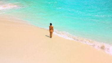 Hava görüntüleri, turkuaz dalgalar içeri girerken parlak bir tropikal kıyı şeridi boyunca ayakta duran bir kadını gösteriyor. Seyahat reklamları, yaz temaları, markalaşma ve yüksek kaliteli plaj yaşam tarzı içeriği için ideal..