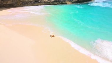 Hava görüntüleri, turkuaz dalgaların Kelingking Sahili, Nusa Penida, Bali 'de yumuşak kumla buluştuğu, seyahat reklamları, sağlık markaları, sosyal medya başlıkları ve plaj kavramları için ideal bir yerde dinlenen bir kadını gösteriyor..