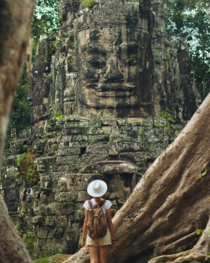 Beyaz şapkalı kadın sırt çantalı dev ağaç kökleri arasında duruyor, Angkor Thom 'daki bir Bayon kulesine bakıyor, Siem Reap, Kamboçya' daki. Turizm reklamları, blogları ve rehberleri için yüksek kaliteli seyahat görüntüsü.
