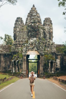 Sırt çantası ve şapkası olan yalnız bir bayan gezgin Kamboçya 'daki antik bir tapınak kapısına doğru yürüyor, yüksek kaliteli promosyon için kültürel miras ve seyahat sahnesi yaratıyor..