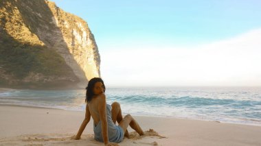 Female traveler at Kelingking Beach on Nusa Penida, Bali, seated on smooth sand with gentle turquoise waves and dramatic limestone cliffs. High quality, conceptual lifestyle photo for ads, blogs, and