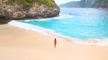 Travel lifestyle image of a woman on Kelingking Beach, Nusa Penida, Bali, standing at the water s edge with golden sand, turquoise surf and towering limestone cliffs. High quality conceptual shot for