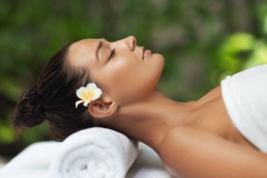 Parlak tenli ve saçlarında tropikal çiçekle lüks kaplıcalarda masaj masasında dinlenen güzel bir genç kadın. Refah, güzellik, cilt bakımı, tatil ve rahatlama için yüksek kaliteli kavramsal görüntüler