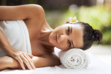 Tropikal kaplıcada rahat bir Asyalı kadın saçı başı altında çiçek ve havluyla uzanmış, nazikçe gülümsüyor. Güzellik kaplıcası ve lüks markalaşma için yüksek kalite sağlık ve cilt bakımı konsepti.