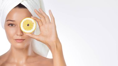 C vitamini konsepti ve reklamı: Havlu türbanlı genç kadın beyaz arka planda bir gözünün üzerinde taze bir limon dilimi tutuyor. Temiz stüdyo portresi, reklamlar için yüksek kalite güzellik fotoğrafı..