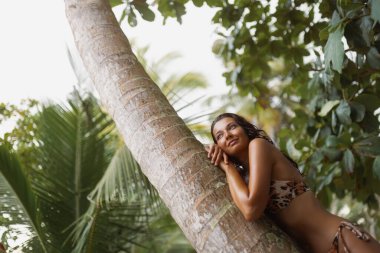 Leopar bikinili güzel bir kadın tropikal bir ortamda palmiye ağacında yatarken gülümsüyor. Yaşam tarzı blogları, egzotik tatil görselleri, doğa inzivaları ve sağlık promosyonları için harika..