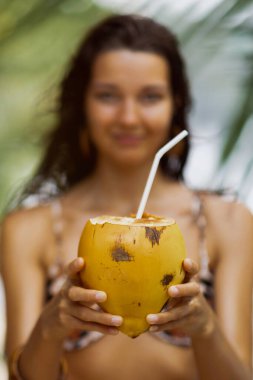 Sarı hindistan cevizi, arka planı bulanık, bikinili bir kadın tarafından ileri uzatılıyor. Sağlık, detoks, tropikal beslenme, plaj suyu ve sağlıklı yaşayan markalar için mükemmel..