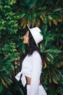 Beyaz şapkalı beyaz bir kadının tropikal portresi. Yeşilliğin yanında duruyor. Moda reklamları, yaşam tarzı markalaşması, seyahat görselleri için ideal yüksek kalite kavramsal promosyon imajı