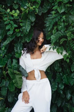 Beyaz yaz kıyafetli zarif beyaz kadın yoğun tropikal yaprakların önünde poz veriyor. Moda tanıtımları, yaşam tarzı kampanyaları, seyahat markaları, tatil kıyafetleri için ideal yüksek kalite kavramsal imaj.