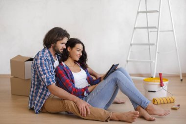 Keyifli bir çift, gündelik giysiler içinde yerde oturmuş, bir tablet kullanarak ev dekorasyonu projesini planlıyorlar. Ev yenileme, iç tasarım ve proje yönetimi kavramları için mükemmel..