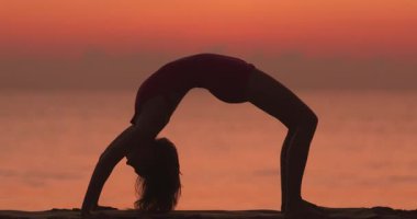 Bir kadın tek bacaklı köprü pozunu gün batımında kumlu bir plajda, arkada okyanusla pratik yapıyor. Esneklik, farkındalık, refah ve huzur kavramları için idealdir.