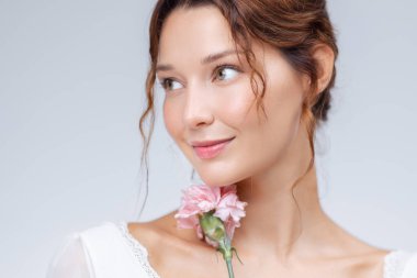 Beyaz tenli ve çenesinin altında pembe karanfil çiçeği olan beyaz bir kadının zarif stüdyo portresi. Cilt bakımı, kozmetik ve rafine güzellik ya da sağlık promosyonları için idealdir..