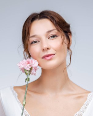 Pembe bir karanfil tutan beyaz tenli genç bir kadının doğal yakın plan portresi. Temiz stüdyo ışığı altında tasarlanmış. Cilt bakımı, çiçek ve güzellik terfileri için mükemmel..