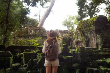 Sırt çantalı ve şapkalı kadın gezgin gür ormanlarda yosun kaplı Angkor taş kalıntıları, seyahat blogları, kültürel turizm kampanyaları, tarih hikayeleri ve macera yaşam tarzı için ideal.