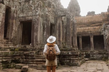 Sırt çantası ve şapka ile Bayon Tapınağı avlusu gezgini Angkor Thom, Kamboçya 'daki antik Khmer taş kalıntıları arasında duruyor. Seyahat blogları, macera turizmi reklamları ve seyahat kampanyaları için ideal..