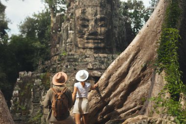 Modern mezar yağmacıları gibi giyinmiş şık bir çift Angkor orman harabelerinde taş kapılar ardında duruyor. Macera kampanyaları, film posterleri, oyun konseptleri ve fantezi hikayeleri için ideal..