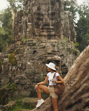 Zengin orman harabelerinde ikonik Bayon taş yüzünün yanında duran kadın turist, macera tanıtımları, kültür turizmi kampanyaları ve yüksek kaliteli editoryal kullanım için mükemmel kavramsal seyahat reklamı imajı.