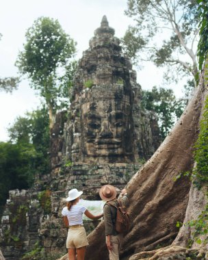 Birkaç genç hazine avcısı harita üzerinde çalışıyor ve bereketli ormandaki Angkor Tapınağı 'nın dev taş yüzlü kapısını gösteriyor. Macera reklamları, fantezi hikayeleri, oyun posterleri ve turizm blogları için ideal.