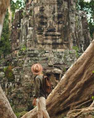 Kamboçya 'daki Angkor Bayon tapınağı, dev ağaç kökleri tarafından çerçevelenmiş, heybetli taş bir yüze hayran olan bir sırt çantalı. Turizm reklamları, seyahat blogları, başyazılar ve macera kullanımı için yüksek kalite, kavramsal imaj.