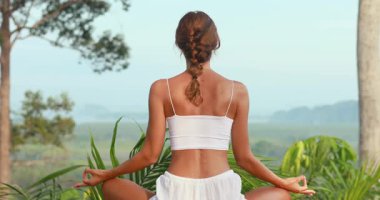Beyaz kıyafetli bir kadının, yemyeşil tropikal bir çevrede tahta bir güvertede bacak bacak bacak üstüne atarak meditasyon yapışının arka görüntüsü. Yoga, akıl sağlığı, dinlenme ve dinlenme reklamları için kavramsal tanıtım..
