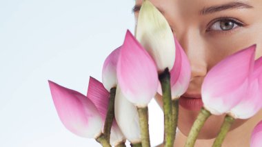 Yüzünün önünde pembe nilüfer çiçekleri tutan bir kadının güzelliğe ve doğal cilt bakımına odaklandığı yakın plan. Güzellik, sağlık ve kaplıca kavramları ya da çiçek temalı reklamlar için ideal..