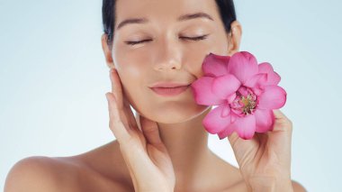 Kapalı gözlü, yüzüne dokunan ve elinde nilüfer çiçeği tutan genç bir kadının yakın plan portresi. Spa, sağlık, kozmetik ve tanıtım konseptleri için yüksek kaliteli güzellik ve cilt bakımı stüdyo görüntüsü.