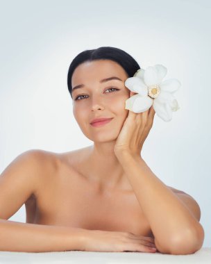 Güzel genç bir kadın yüzünün yanında beyaz bir çiçekle poz verir, doğal güzelliği ve sağlığı sembolize eder. Çıplak omuzları ve sakin ifadesi, cilt bakımı için ideal tazeliği ve zarafeti vurguluyor.