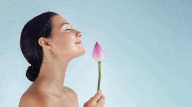 Gülümseyen bir kadının profili pembe nilüfer tomurcuğunu nazikçe tutuyor, gözleri kapalı, fotokopi alanı olan yumuşak mavi bir arka plana karşı. Sağlık reklamları, cilt bakımı ve spa markaları için ideal..