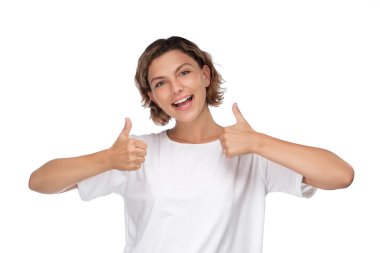 Kısa kıvırcık saçlı genç bir kadın, beyaz bir arka planda izole edilmiş, parlak bir gülümsemeyle çifte onay veriyor. Olumlu temalar, onay ve coşku için ideal.