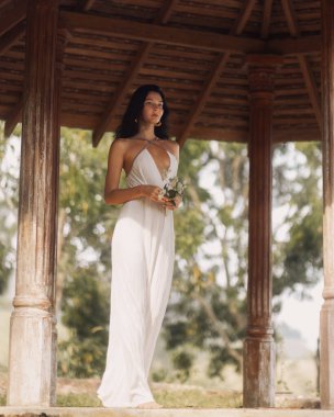 Dalgalı beyaz elbiseli zarif genç bir kadın kırsal bir tropikal çardakta çiçeklerle çıplak ayakla duruyor. Reklamlar ve editoryal kullanım için mükemmel yüksek kaliteli yaşam tarzı, moda veya gelin konsepti.