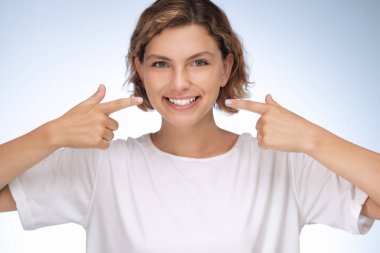 Işıl ışıl gülümseyen neşeli genç bir kadın açık mavi arka planda beyaz dişlerine işaret ediyor, diş sağlığı, güven ve pozitiflik temalarını vurguluyor. Diş reklamları için ideal.