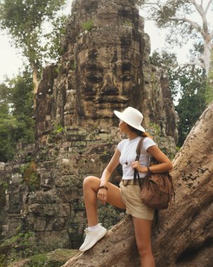 Kamboçya 'da antik Bayon taşından bir yüzün yanında poz veren genç kadın turist, konsept seyahat promosyonu imajı macera turizmi için ideal, yüksek kaliteli editoryal tasarımlar ve ilham verici kültürel keşif içeriği