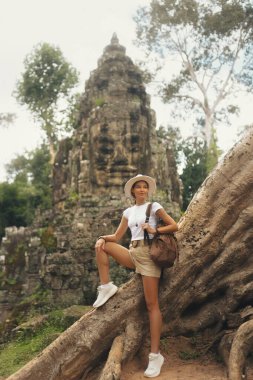 Gülen kadın turist Kamboçya 'nın ormanında büyük bir Bayon taşından yüzle dinleniyor, kavramsal seyahat reklamları yüksek kaliteli turizm tanıtımları, macera kampanyaları ve editoryal görseller için mükemmel.