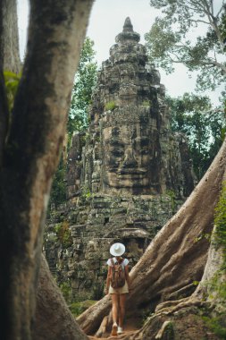 Beyaz şapkalı Angkor Thom sırt çantalı dev ağaç kökleri arasında duruyor, yemyeşil orman harabelerinde bir Bayon yüz kulesine bakıyor. Konsept, seyahat reklamları, bloglar, sosyal paylaşımlar ve turizm rehberleri için yüksek kaliteli görüntü