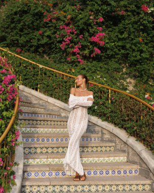 Beyaz dantel elbiseli genç bir kadın şık bir şekilde Sunset Town, Phu Quoc 'taki renkli ejder merdiveninde poz veriyor. Etrafı yemyeşil çiçeklerle çevrili romantik ve ikonik bir seyahat noktası..
