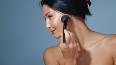 Pürüzsüz, parlak bir cilde sahip fırçayla allık süren bir kadının yakın plan portresi. Cilt bakımı, kozmetik, makyaj dersleri ya da temiz mavi arka planda sağlığın tanıtımı için mükemmel..