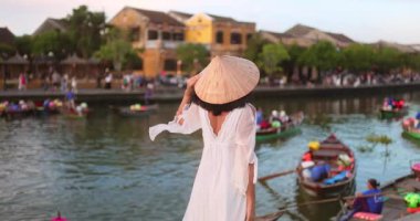 Beyaz elbiseli ve konik şapkalı şık Asyalı kadın Hoi An, Vietnam 'da nehir kenarında duruyor. Kavramsal seyahat tanıtımı, turizm, moda, yaşam tarzı ve kültür reklamları için ideal..