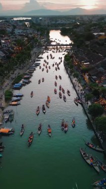 Hoi An nehri üzerinde gün batımında düzinelerce fener teknesi kalabalık bir köprüye doğru sürüklenirken, suya yansıyan yansımalar. Günümüzde seyahat reklamları, turizm tanıtımları ve Vietnam kültür temaları için ideal..