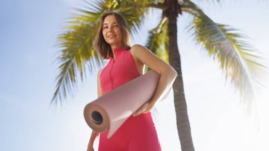Parlak pembe elbiseli gülümseyen bir kadın dışarıda palmiye ağaçlarının altında yoga minderi tutuyor. Tropikal manzara ve onun neşeli ifadesi sağlıklı yaşam ve zindelik için mükemmel bir imaj yaratıyor..