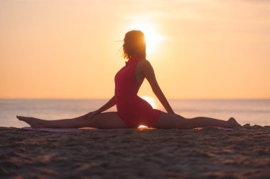 Pembe aktif giysili bir kadın silueti gün batımında bir plajda sicim yoga pozu veriyor. Parlayan güneş ve sakin okyanus, refahı, rahatlığı ve rahatlığı arttırmak için ideal bir zemin sağlar.