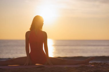 Silhouetted woman in pink activewear performs a full split on a yoga mat at the beach during sunrise. She embraces mindfulness, flexibility, and relaxation in a peaceful outdoor setting.