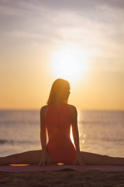 Kırmızı elbiseli atletik bir kadın güneş doğarken kumlu sahilde yoga yapıyor. Parlayan güneş tarafından silueti var. Sıhhat, esneklik, ruhsal dinlenme ve vücut bakımı temaları için ideal..