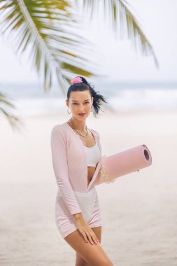 Kendine güvenen, beyaz spor bluzlu, şortlu, pembe hırkalı ve plajda yoga minderiyle altın zincirli seksi Asyalı kadın. Yüksek kaliteli yaşam tarzı, moda ve tanıtım amaçlı sağlık portresi konsepti