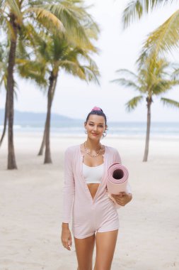 Pembe elbiseli kendine güvenen bir kadın tropik bir plajda yoga minderi taşıyarak yürüyor. Yaşam tarzı, fitness ve sağlık temaları için mükemmel, sergilenen rahatlama, sağlık, ve modaya uygun atletizm