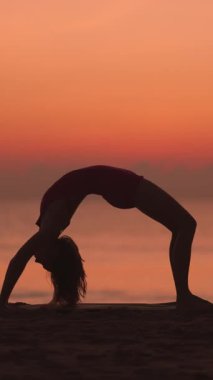 Bir kadın gün doğumunda plajda tekerlek pozu veriyor, gökyüzüne karşı silueti var. Bu barışçıl, yavaş çekim sahnesi esnekliği, gücü ve farkındalığı vurguluyor. Formda ve sağlıklı olmak için mükemmel.
