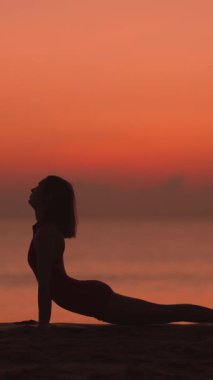 Dikey videoda, bir kadın gün batımında plajda yoga yapıyor, poz verirken siluette yakalanmış. Canlı gökyüzü ve sakin okyanus dinginliği arttırır ve sağlığa odaklanır.