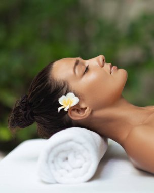 Masaj masasında dinlenen Spa 'lı kadın sarılan havlu ve Plumeria çiçeğiyle, gözleri huzur içinde kapalı. Cilt bakımı reklamları, güzellik salonları, otel spa promosyonları ve kişisel bakım için ideal..