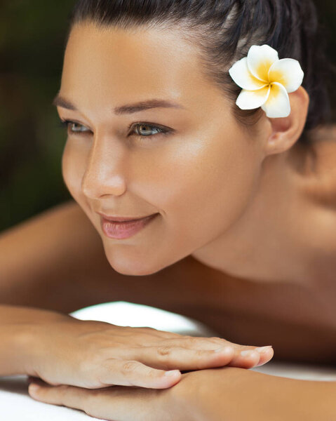Caucasian woman with glowing skin and a frangipani flower in her hair smiles softly while resting at a spa. Perfect for wellness, skincare, relaxation, and luxury beauty promotions.