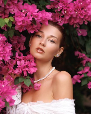 Dantel bir elbise ve inci takan zarif, genç beyaz bir kadın, yemyeşil begonviller içinde poz veriyor. Stok kullanımı için yüksek kaliteli moda, güzellik veya çiçek tanıtım konsepti.