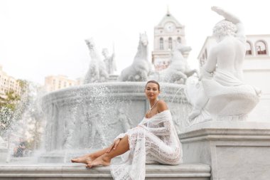 Beyaz dantel elbiseli ve incili zarif kadın Phu Quoc, Vietnam 'daki Çeşme At' ın mermer çıkıntısında yalınayak oturuyor. Yayın ve reklamlar için yüksek kalite moda, seyahat veya gelin konsepti.